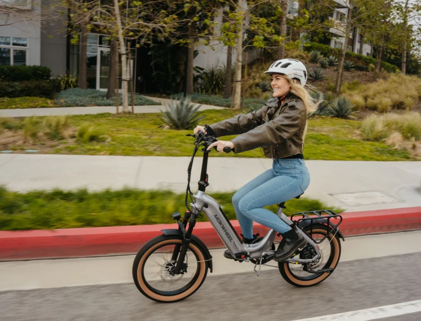 A woman wearing a helmet and jacket rides an electric bicycle on a street next to a sidewalk bordered by greenery.