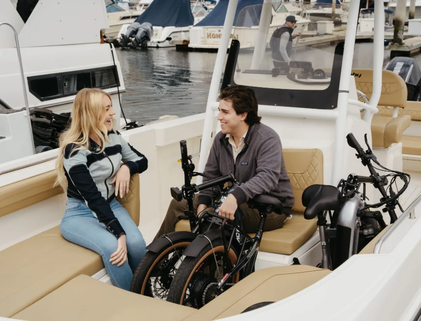 A woman and a man sit on a boat facing each other, talking and smiling, with two folded electric bikes between them and other boats visible in the background.
