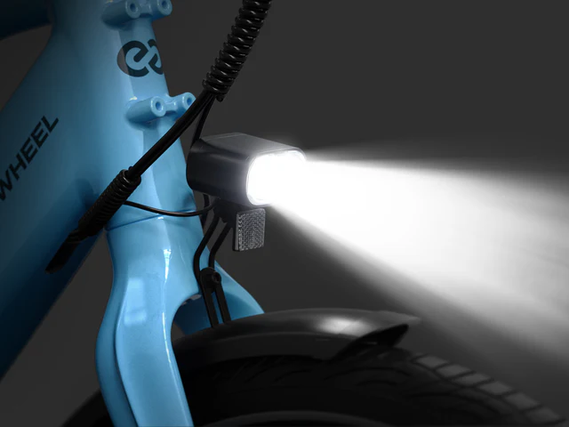 Close-up of a bicycle front light turned on, mounted above the front wheel of a blue bike, illuminating a bright beam forward.