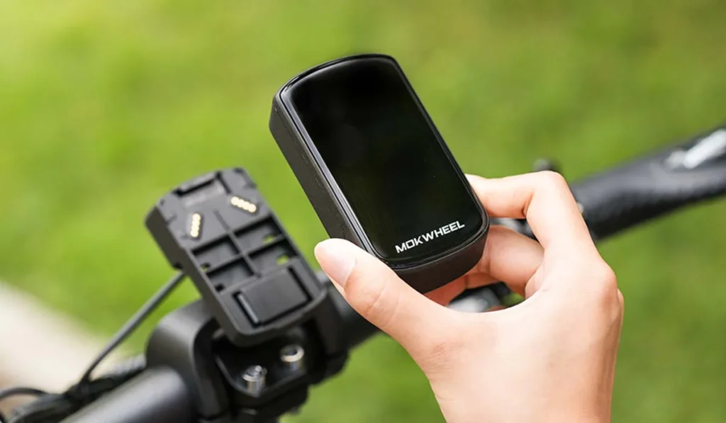 A person holds a MOKWHEEL device above its mounting bracket on a bicycle handlebar, with a grassy background visible.