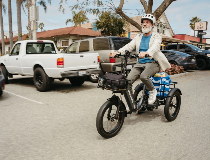 An older man wearing a helmet and blazer rides an electric tricycle on a street, carrying bags in the rear basket. Cars and trees are visible in the background.
