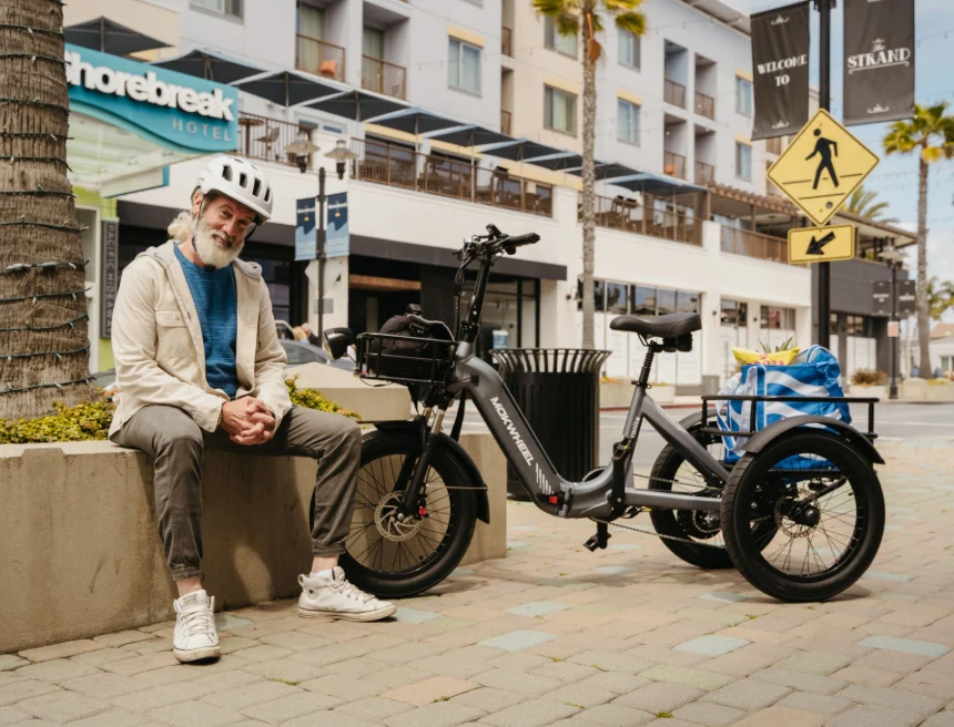 An older man in casual clothes and a helmet sits on a low wall next to an electric tricycle with shopping bags in an urban outdoor setting.