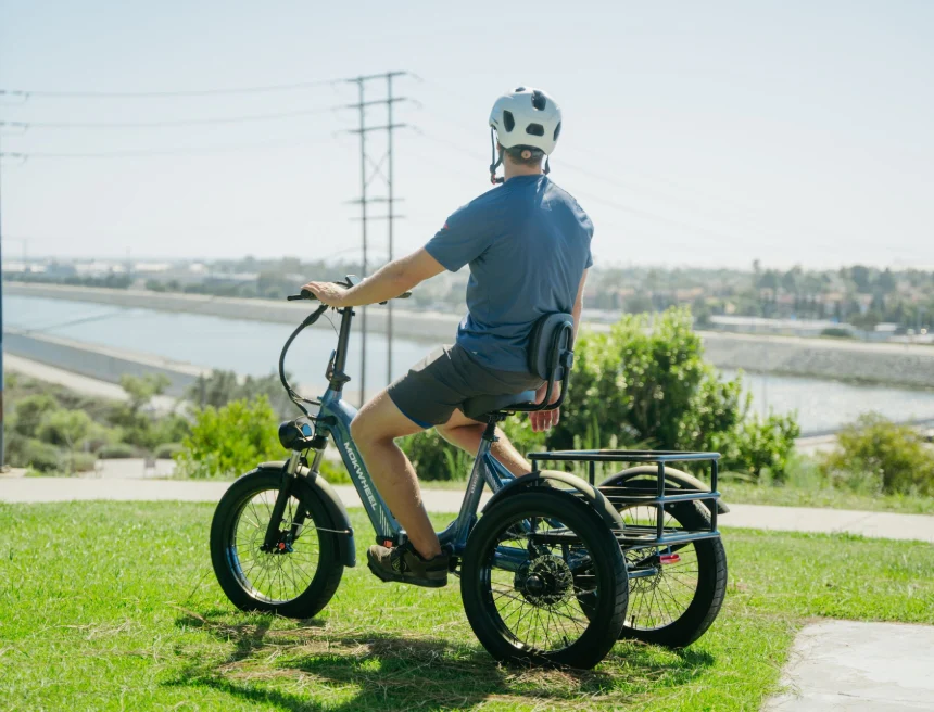 A person wearing a helmet sits on an electric tricycle, facing away and overlooking a river and cityscape on a sunny day.