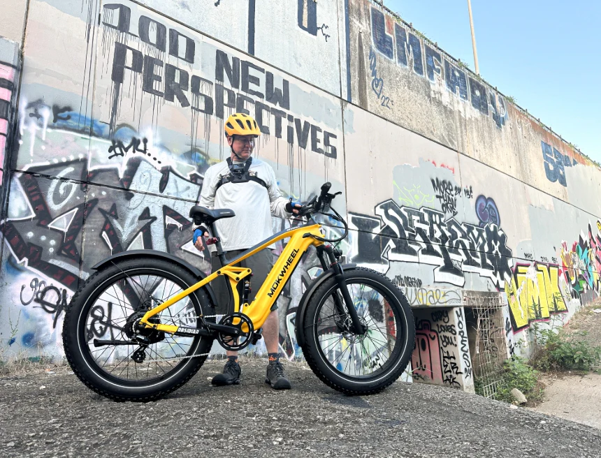 A person in cycling gear and helmet stands beside a yellow e-bike in front of a graffiti-covered wall that reads "100 NEW PERSPECTIVES.