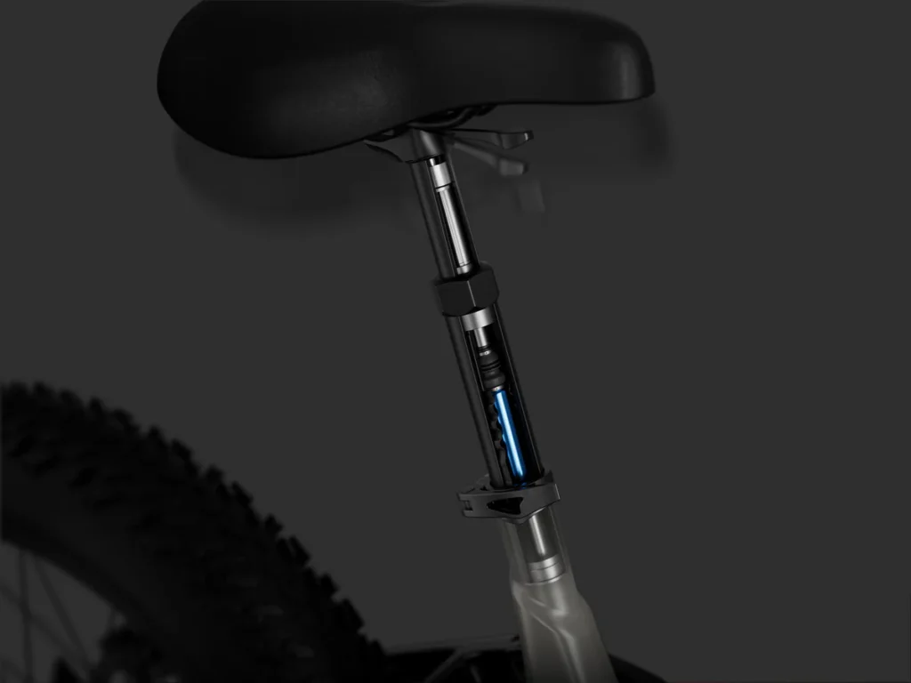 Close-up of a bicycle seat post with an exposed internal mechanism, showing part of the bike frame and rear tire against a dark background.