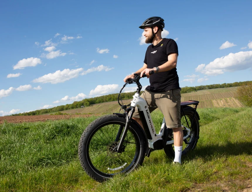 A person wearing a helmet stands outdoors beside an electric bicycle on a grassy path, with open fields and a blue sky in the background.