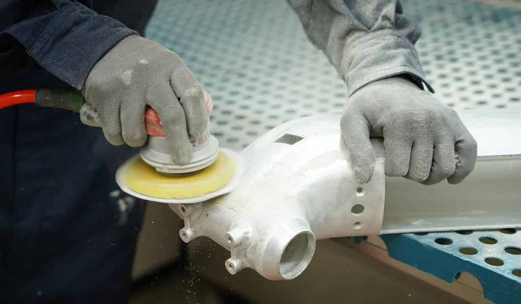 A person wearing gloves uses a power sander to polish a metallic part, with dust particles visible during the sanding process.