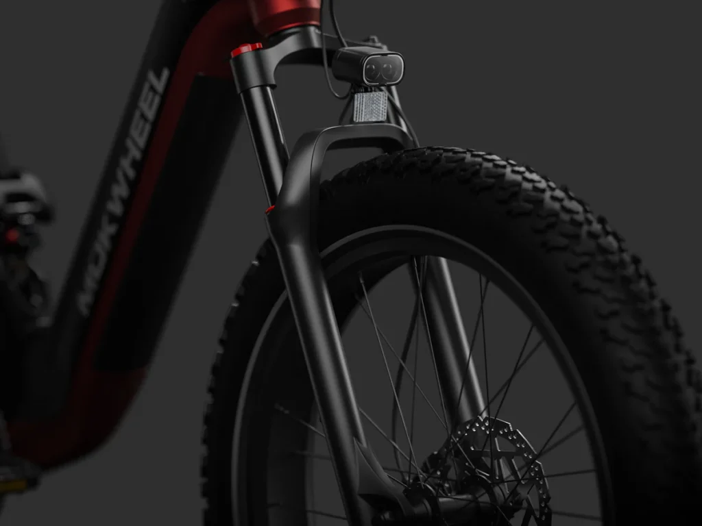Close-up view of the front wheel and fork of a Mokwheel electric bicycle with a fat tire, disc brake, and front-mounted light against a dark background.