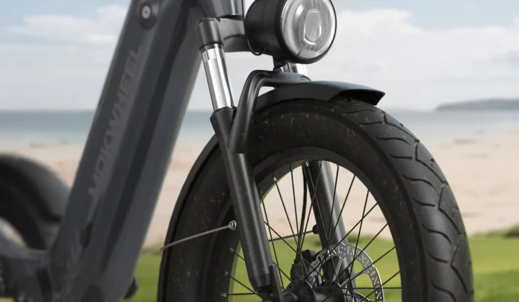 Close-up of the front wheel and suspension fork of an e-bike with disc brakes, parked on grass near a sandy beach with water in the background.