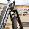Close-up of the front section of a Slate Ebike, showcasing its 80mm suspension fork, with a marina and buildings softly blurred in the background.