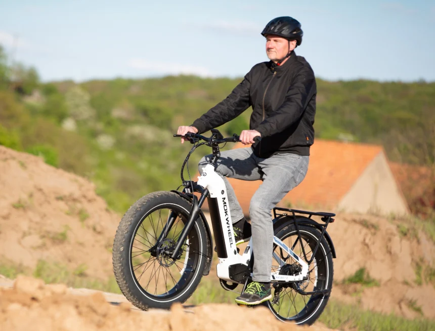 A person wearing a black jacket, gray pants, and a helmet rides a white fat-tire electric bike outdoors on a dirt path with greenery and buildings in the background.