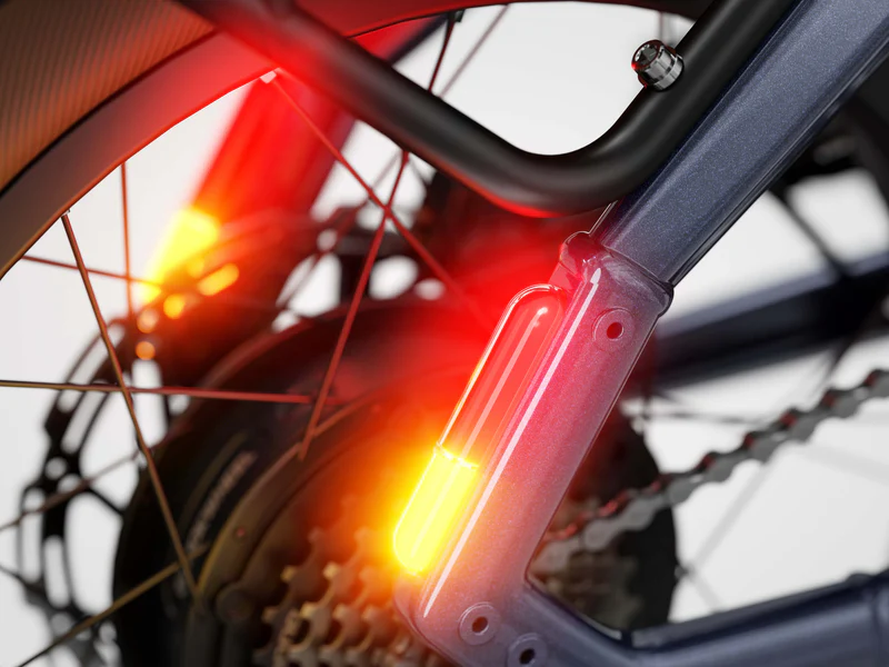 Close-up of a bicycle's rear wheel, showing illuminated yellow and red integrated lights near the frame, spokes, gears, and tire.