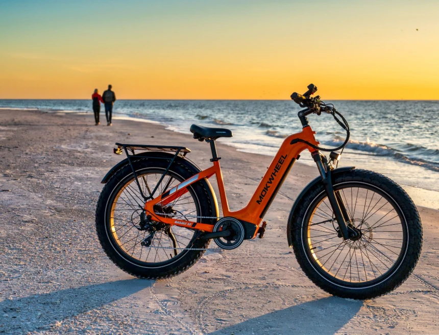 An orange electric fat-tire bike is parked on a sandy beach at sunset, with two people walking near the shoreline in the background.