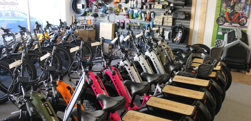 A bike shop interior with rows of electric bicycles lined up, accessories and helmets displayed on shelves, and various bike parts visible in the background.