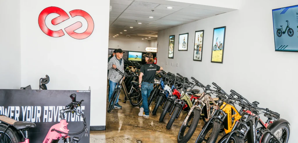 Two people talk in a well-lit bike shop lined with electric bikes, posters on the wall, and a red infinity-like logo displayed above the entrance.