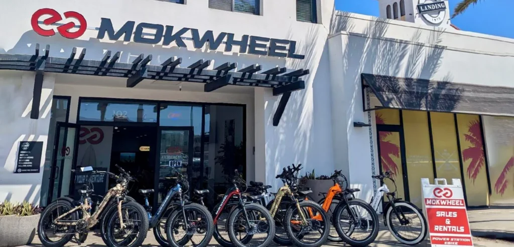A row of electric bikes is parked outside a Mokwheel store, with signage indicating sales and rentals, on a sunny day.