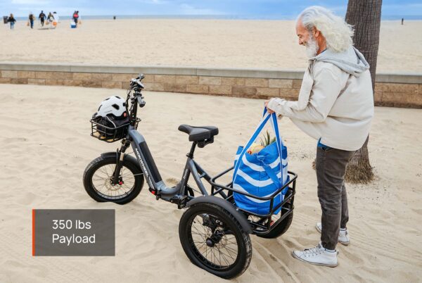 An older man stands on the beach beside the Granite E-Trike, a three-wheeled electric bike featuring a front basket and rear rack with a striped bag. Text: "350 lbs Payload." The ocean is visible in the background.