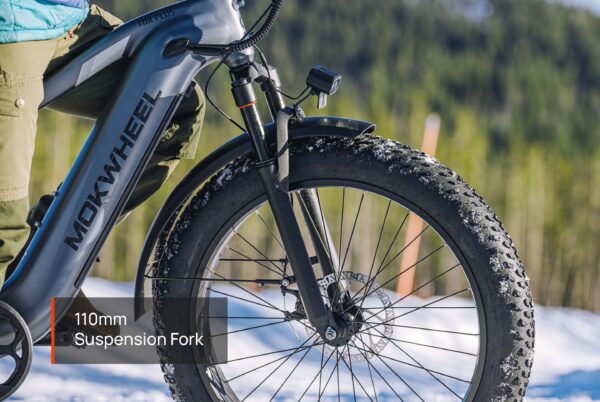 Close-up of the Tor Plus Ebike’s front wheel, featuring a suspension fork marked 110mm, with snowy terrain and trees visible in the background.