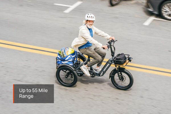 Older adult wearing a helmet rides the Granite E-Trike with bags in the rear basket on a city street. Text overlay reads: “Up to 55 Mile Range.”.