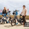 Two people with Slate Ebikes stand on a sandy beach near colorful surfboard-shaped bike racks. Text reads: "Slate Ebike – Up to 60 Mile Range.