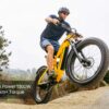 A man rides the Obsidian ST Ebike, a yellow electric bike, up a dirt incline with the front wheel lifted. Text on image: "Peak Power 1300W, 105Nm+ Torque.