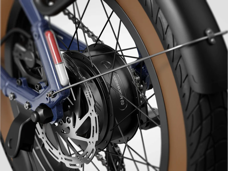 Close-up of a bicycle rear wheel showing a disc brake, a black hub motor labeled "MAXWHEEL," and a brown tire with a black fender.
