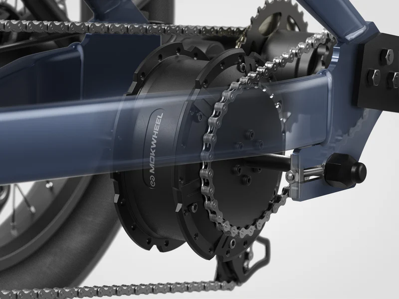 Close-up view of an electric bicycle drivetrain featuring a chain running over gears and a mid-drive motor labeled "Mokwheel.