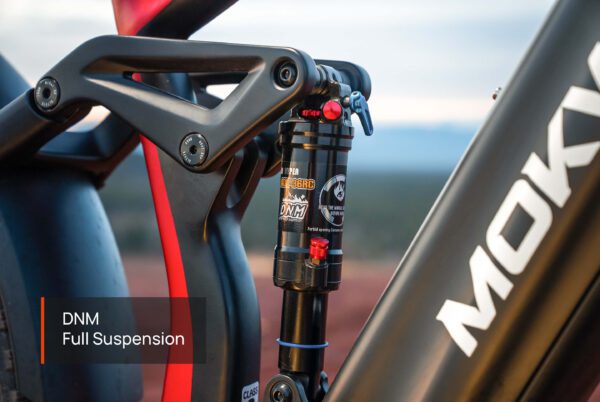 Close-up of a DNM full suspension shock absorber mounted on the black and red frame of the Obsidian ST Ebike, with partial “MOKA” branding visible.