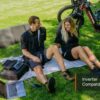 Two people sit on a blanket on grass near two Obsidian Ebikes, a portable solar panel, power station, and laptop. The words "Inverter Compatible" appear in the image.