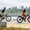 Two people ride Obsidian Ebikes on a paved path by the water, with a caption stating "Up to 80 Mile Range.