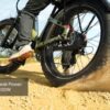 A person rides the Basalt ST 2.0 Ebike across sandy terrain, its fat rear tire spinning and kicking up dust. Text overlay: “Peak Power 1100W.”.