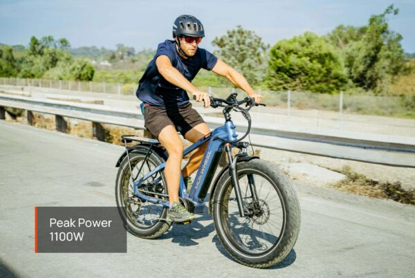 A man in a helmet and sunglasses rides the Basalt Ebike with fat tires on a paved road; a label reads "Peak Power 1100W.