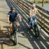 Two people wearing helmets ride Basalt Ebikes across a wooden bridge over water. Text on image reads, "Up to 80 Mile Range.