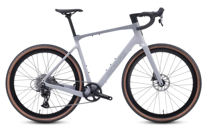 A matte gray gravel bike with drop handlebars, disc brakes, and wide tan-wall tires is shown in a side view against a white background.