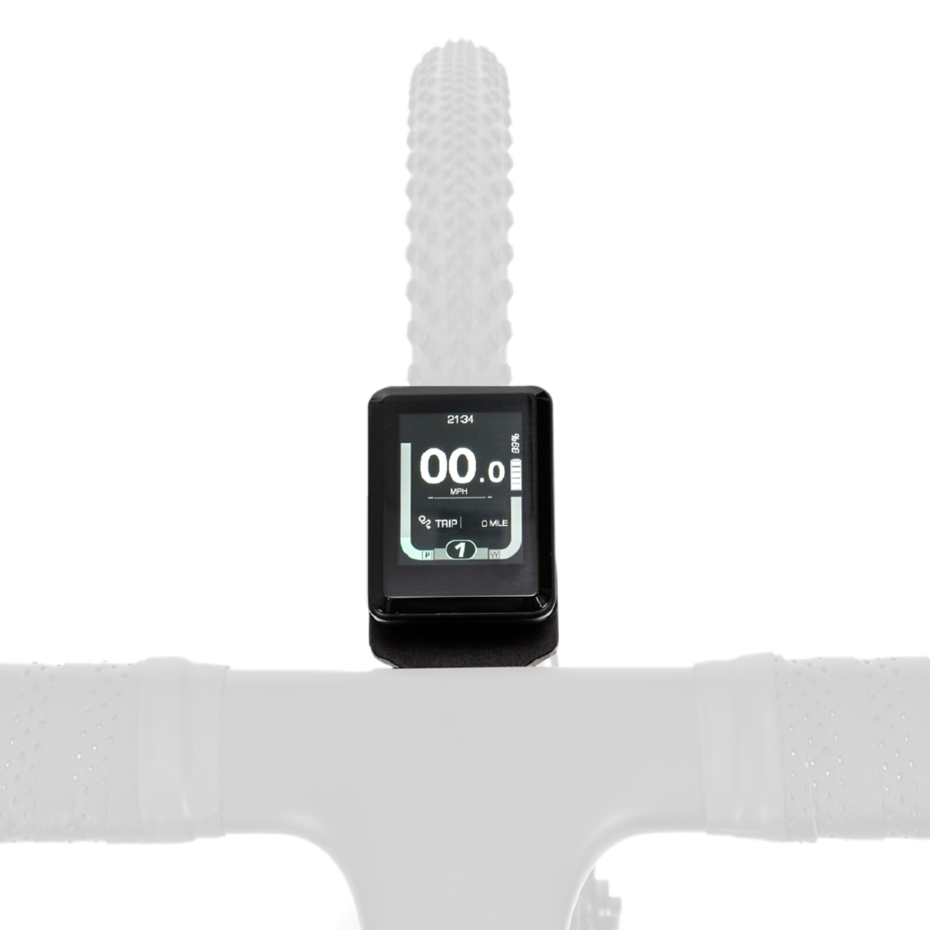 Close-up of a bicycle handlebar with a digital display showing speed, trip distance, and battery level, positioned above the front wheel with a knobby tire.