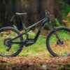 A black full-suspension mountain bike with thick tires is parked on a leaf-covered forest floor with trees and greenery in the background.