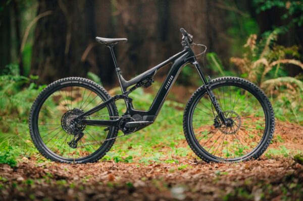 A black full-suspension mountain bike with thick tires is parked on a leaf-covered forest floor with trees and greenery in the background.