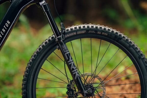 Close-up of a mountain bike's front wheel, suspension fork, and tire, set against a blurred outdoor background with greenery.