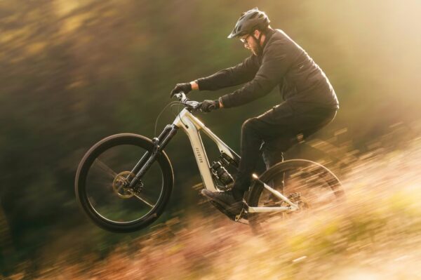 Person wearing a helmet and gloves rides an electric mountain bike on a dirt trail, lifting the front wheel, with blurred background suggesting motion.