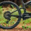 Close-up of the rear wheel and drivetrain of a black mountain bike with Maxxis tires, positioned outdoors on a leafy ground.