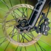 Close-up of a bicycle front wheel showing a disc brake rotor and caliper mounted on a black fork, with a blurred green background.