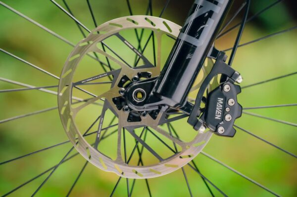 Close-up of a bicycle front wheel showing a disc brake rotor and caliper mounted on a black fork, with a blurred green background.