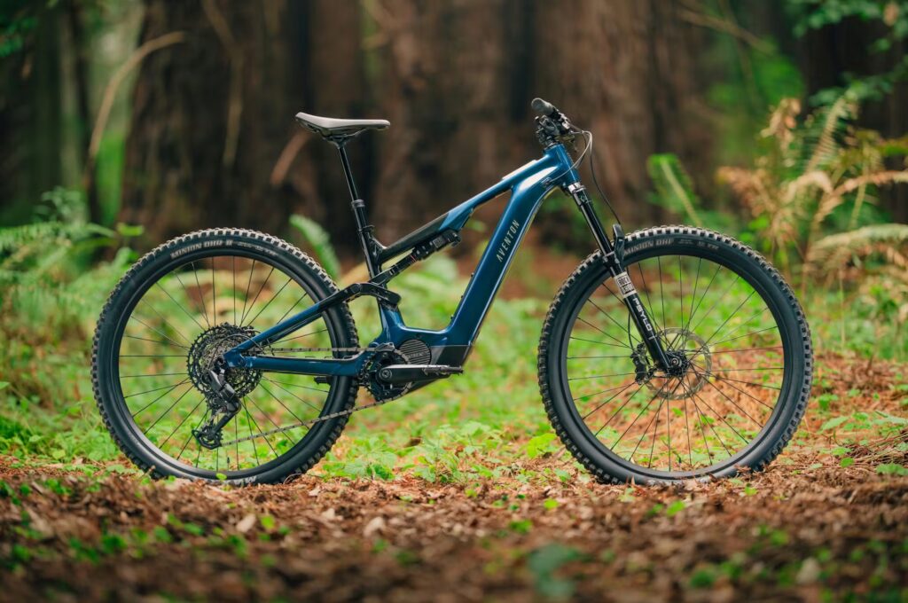 The Current ADV Ebike, a blue full-suspension electric mountain bike, is parked on a leafy forest floor surrounded by trees and greenery.