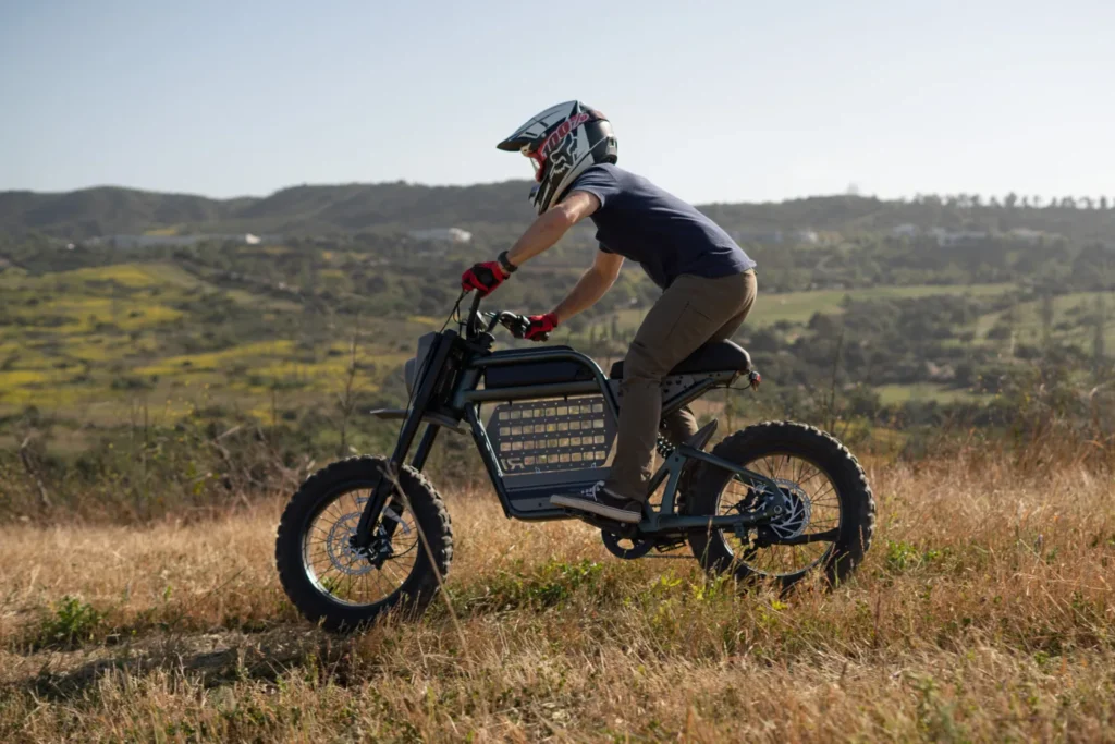 A person wearing a helmet and gloves rides an electric dirt bike across a grassy field with hills in the background, showcasing the thrill of ebikes and essential e-bikes accessories for outdoor adventures.