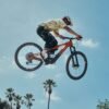 A mountain biker performing a jump in the air against a clear blue sky with palm trees below.