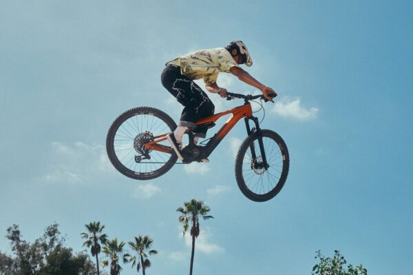 A mountain biker performing a jump in the air against a clear blue sky with palm trees below.
